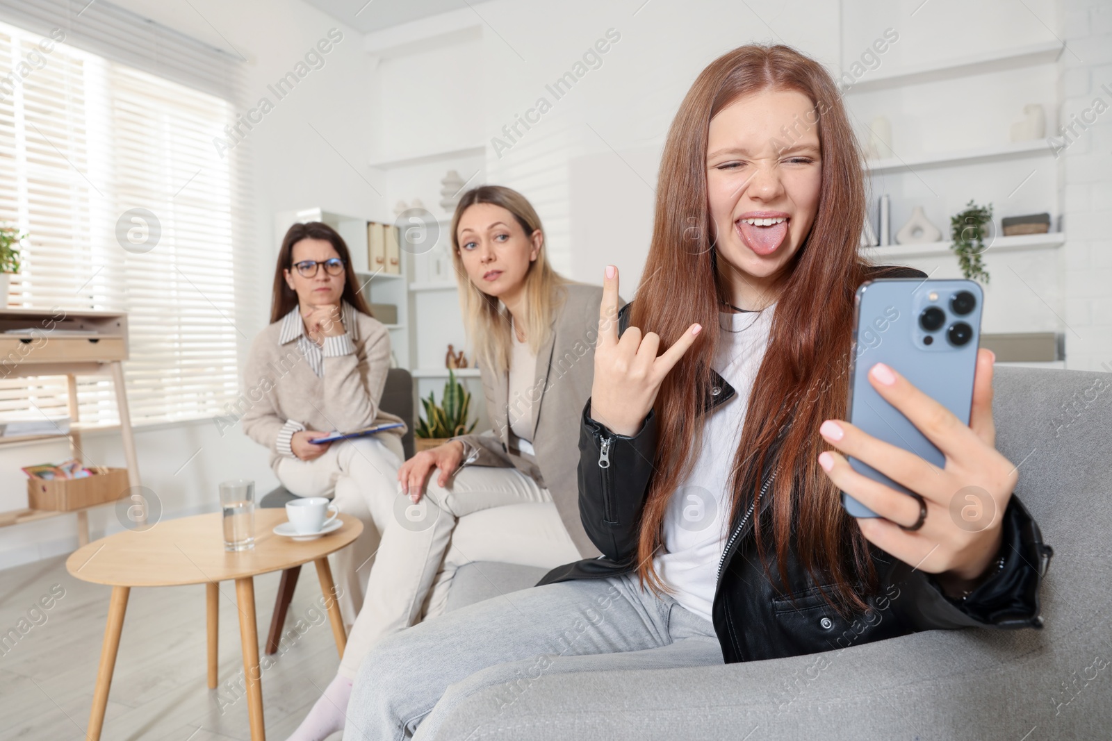 Photo of Rebellious teenage girl filming video during consultation with her mom and psychologist in office, selective focus