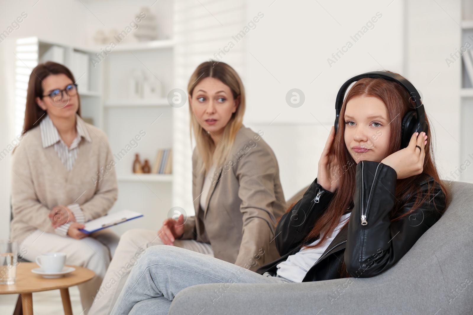 Photo of Annoyed teenage girl listening to music instead of working with psychologist in office, selective focus. Mother and daughter difficult relationship