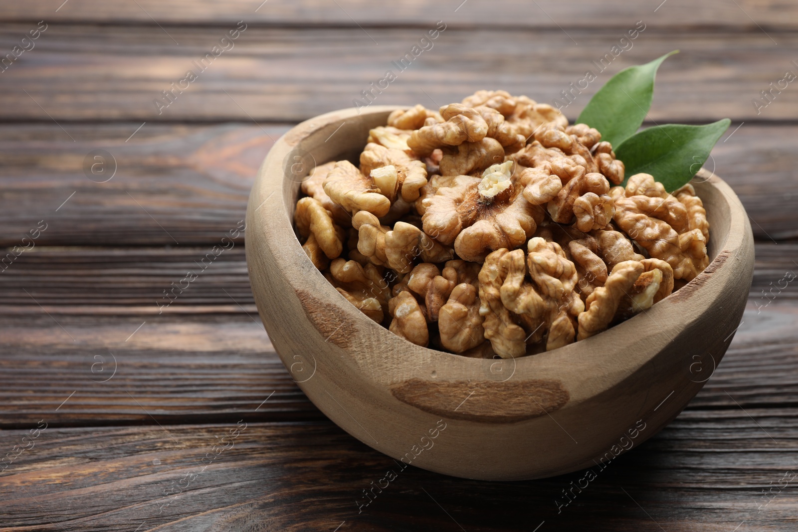 Photo of Peeled walnuts and green leaves in bowl on wooden table, closeup. Space for text