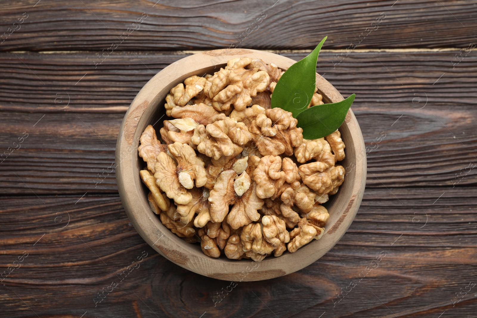 Photo of Peeled walnuts and green leaves in bowl on wooden table, top view