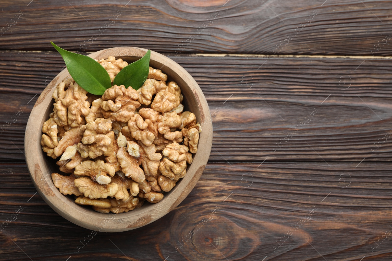 Photo of Peeled walnuts and green leaves in bowl on wooden table, top view. Space for text