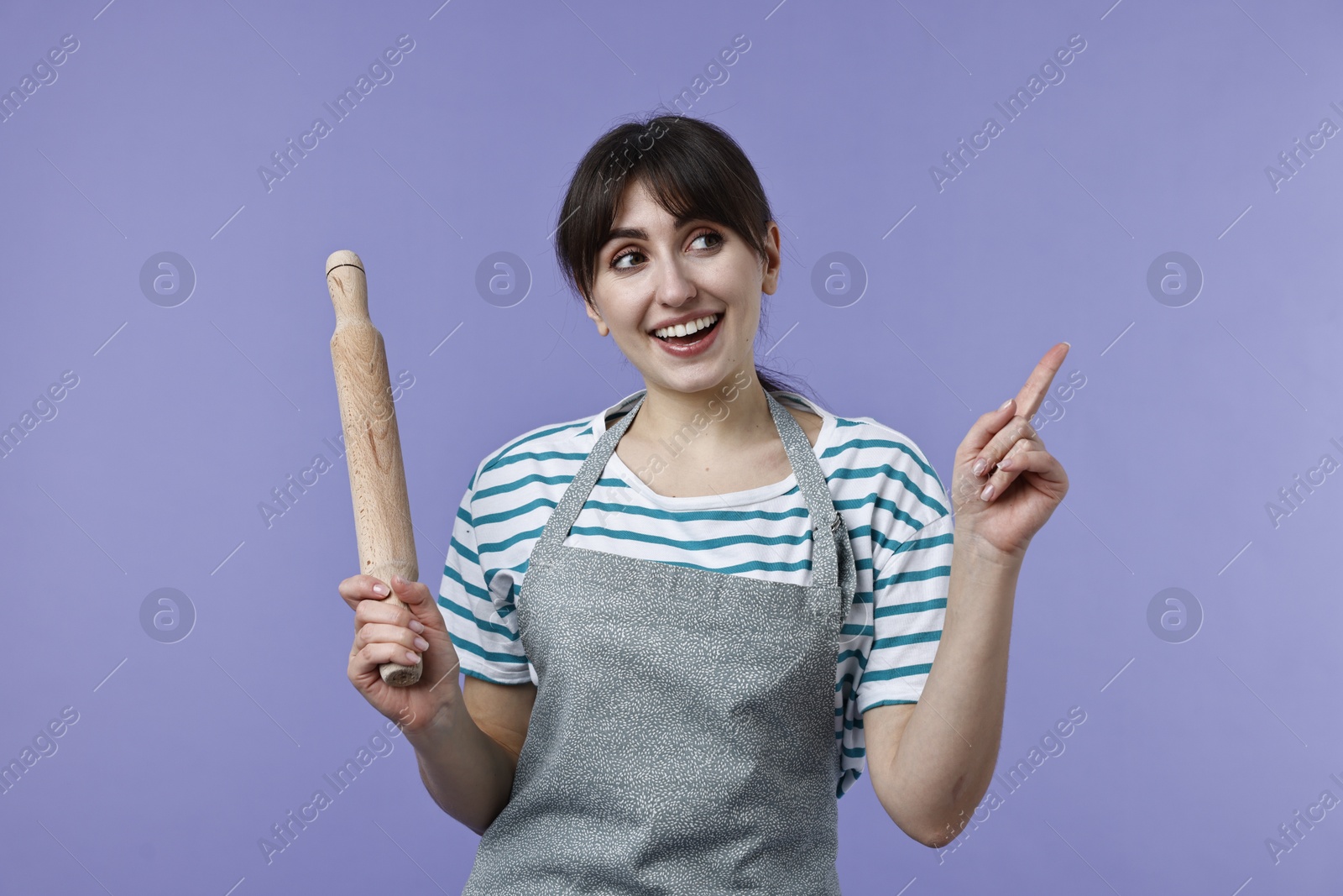 Photo of Happy woman with rolling pin pointing at something on violet background