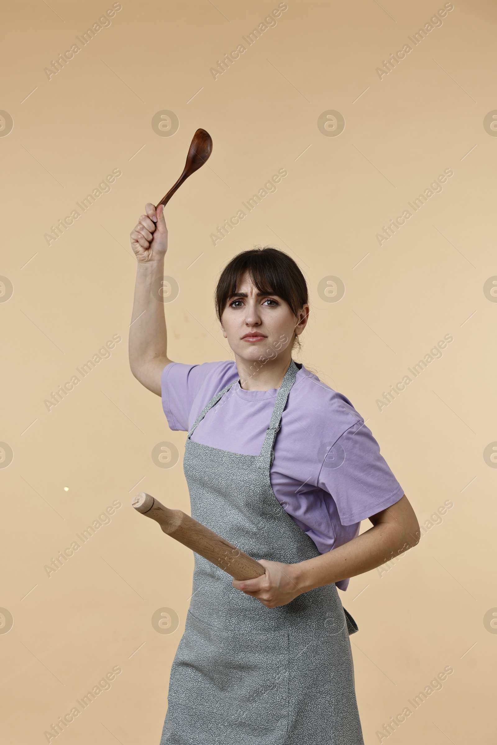 Photo of Angry woman with rolling pin and spoon on beige background