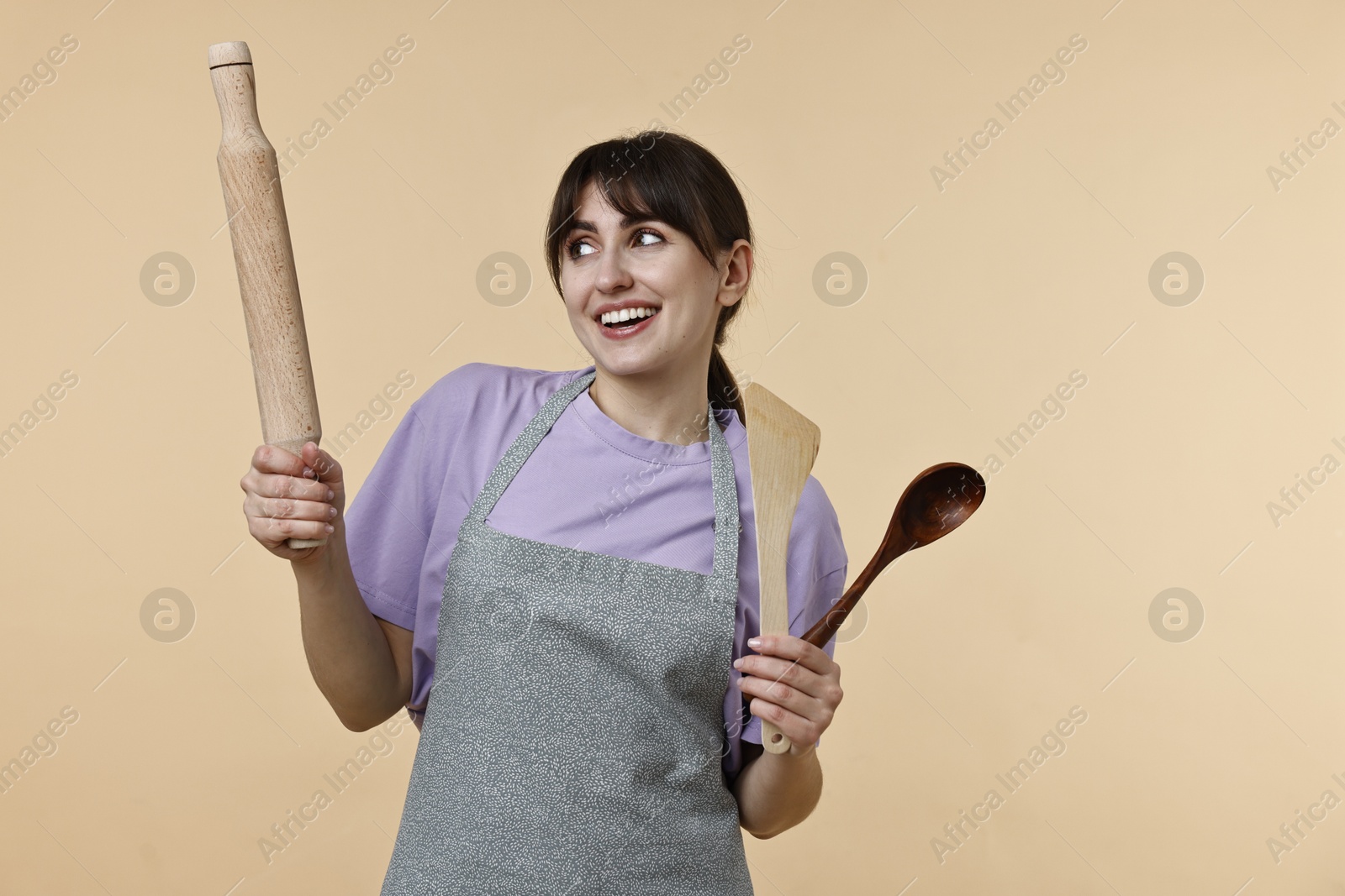 Photo of Happy woman with rolling pin, turner and spoon on beige background