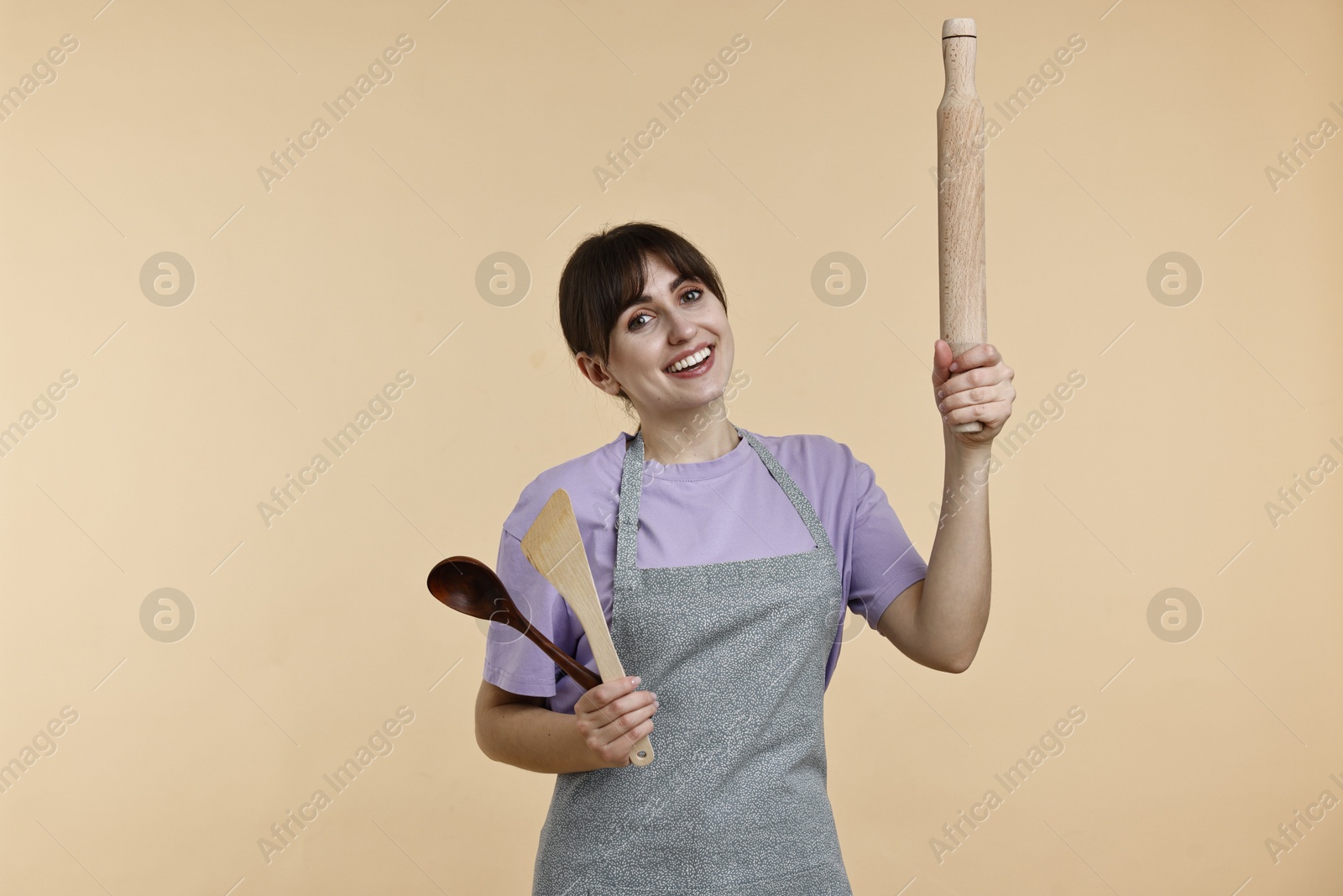 Photo of Happy woman with rolling pin, turner and spoon on beige background