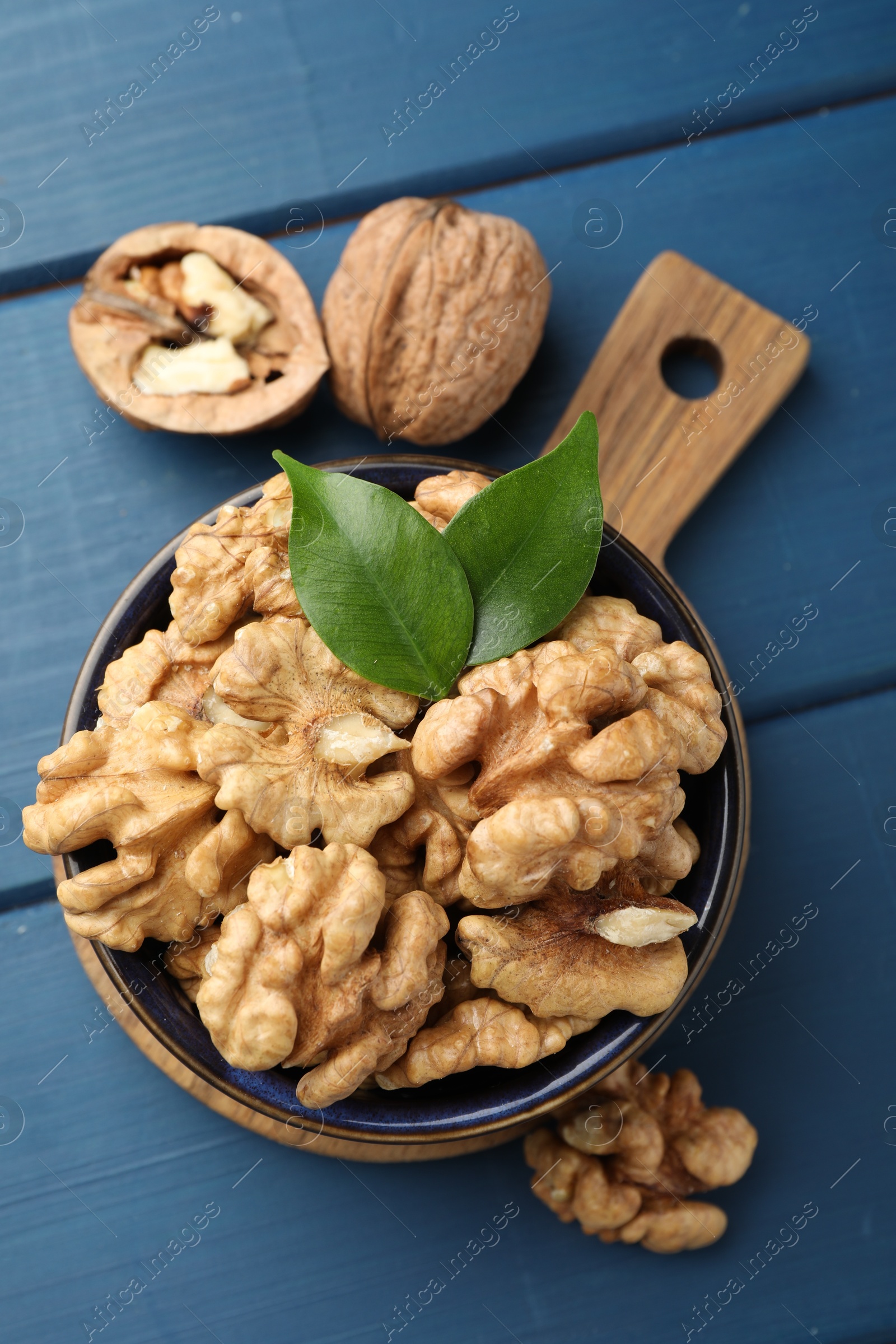 Photo of Fresh walnuts and green leaves in bowl on blue wooden table, flat lay