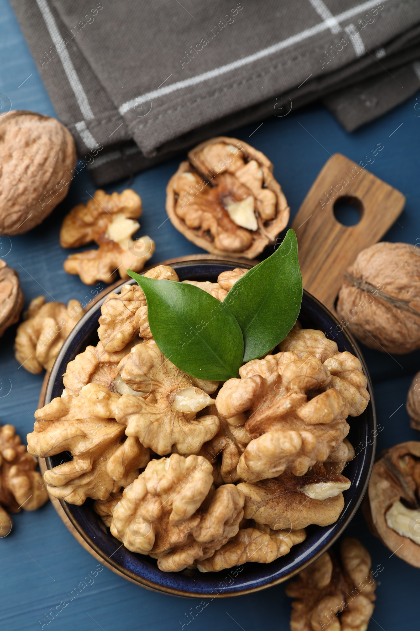 Photo of Fresh walnuts and green leaves in bowl on blue wooden table, flat lay