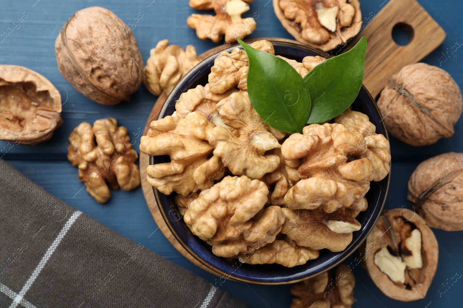 Photo of Fresh walnuts and green leaves in bowl on blue wooden table, flat lay