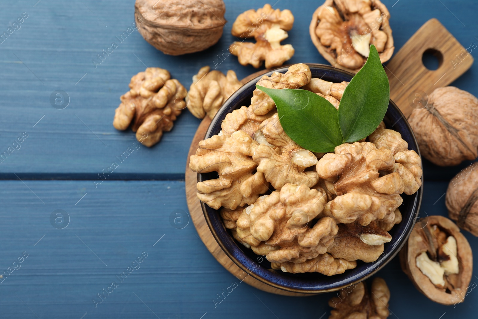 Photo of Fresh walnuts and green leaves in bowl on blue wooden table, flat lay. Space for text