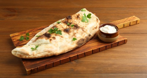 Photo of Board with tasty calzone, parsley and sauce on wooden table, closeup