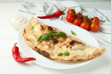 Photo of Tasty calzone, basil, chili pepper, sauce and tomatoes on white wooden table, closeup