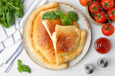 Photo of Pieces of delicious calzone pizza with mozzarella, tomatoes and basil on white tiled table, flat lay