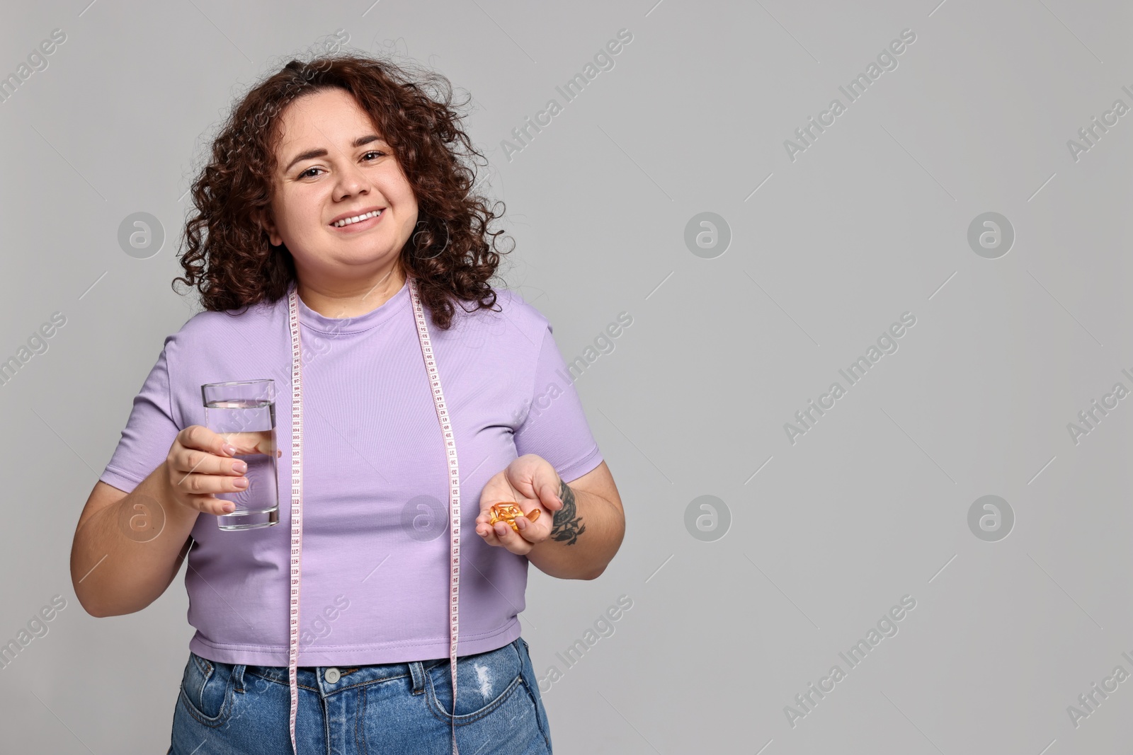 Photo of Happy plus size woman holding pile of weight loss supplements and glass of water on grey background. Space for text