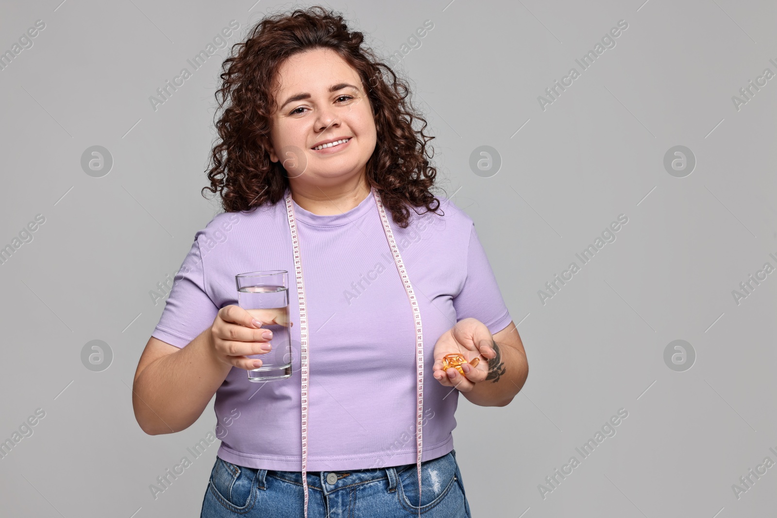 Photo of Happy plus size woman with pile of weight loss supplements and glass of water on grey background