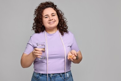 Photo of Happy plus size woman with pile of weight loss supplements and glass of water on grey background