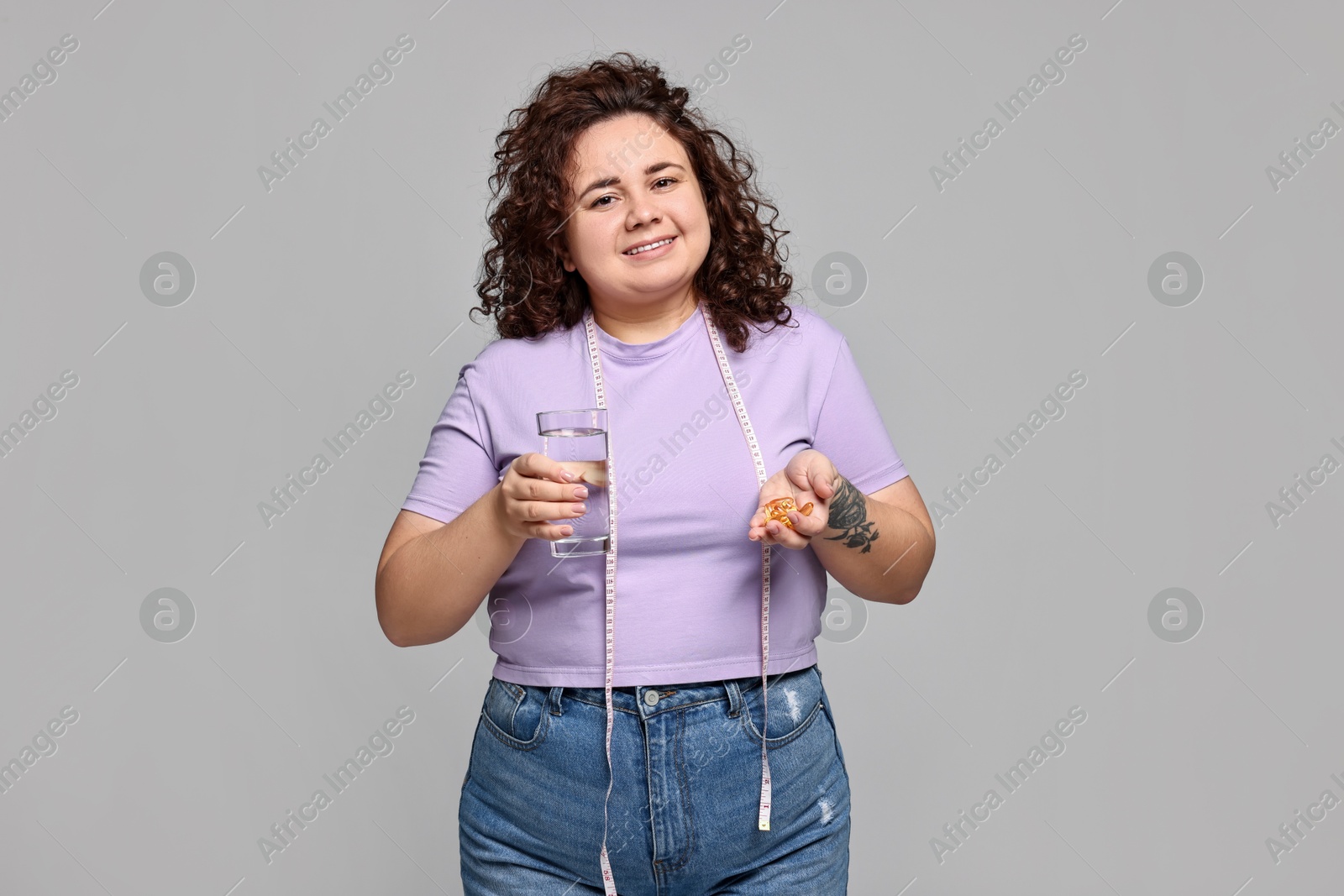 Photo of Happy plus size woman with pile of weight loss supplements and glass of water on grey background