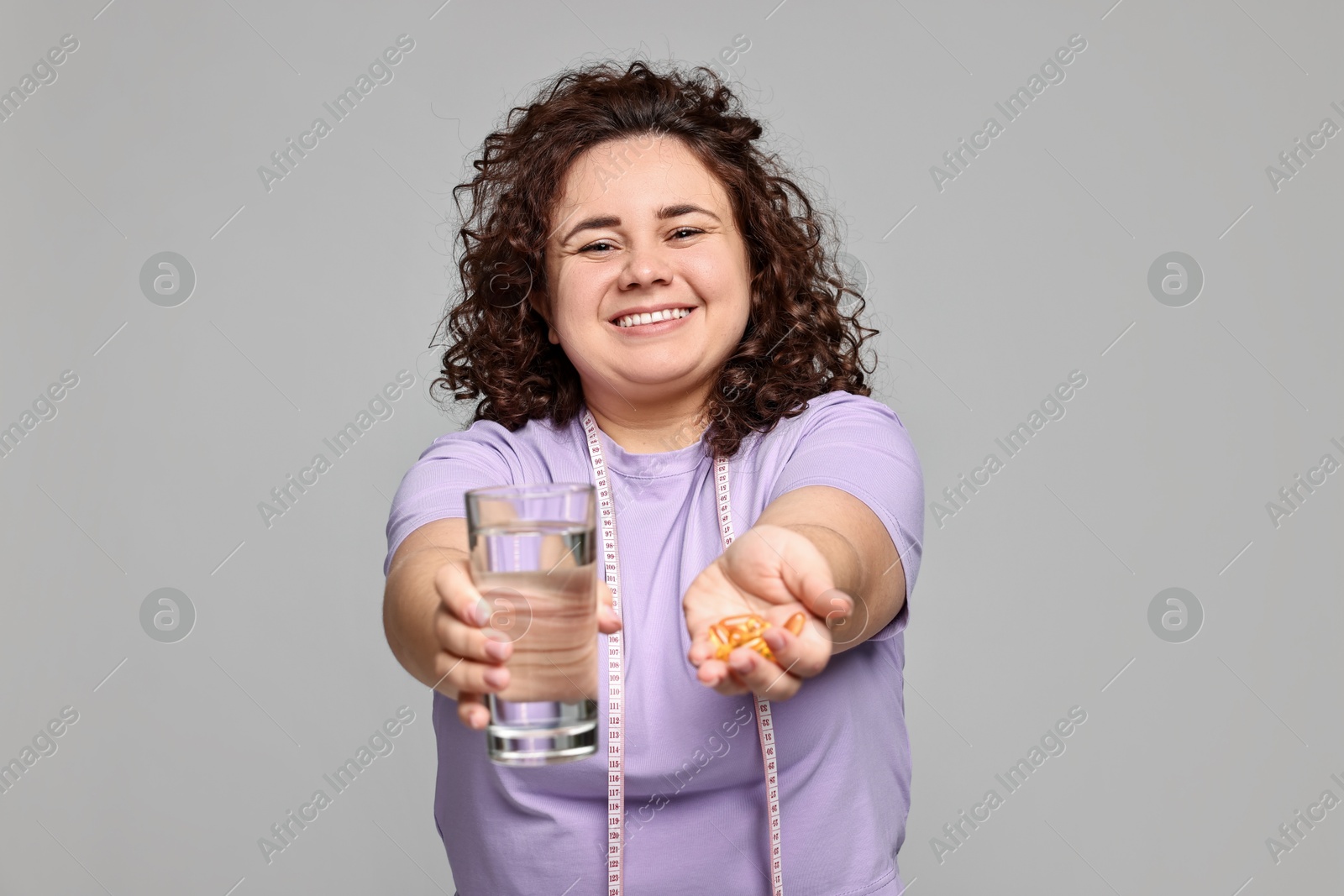 Photo of Happy plus size woman giving pile of weight loss supplements and glass of water on grey background, selective focus