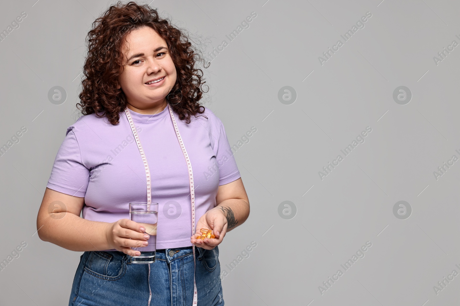 Photo of Happy plus size woman with pile of weight loss supplements and glass of water on grey background. Space for text