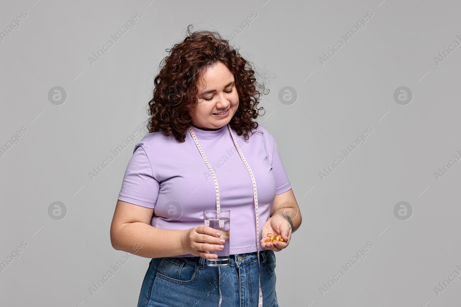 Photo of Happy plus size woman with pile of weight loss supplements and glass of water on grey background