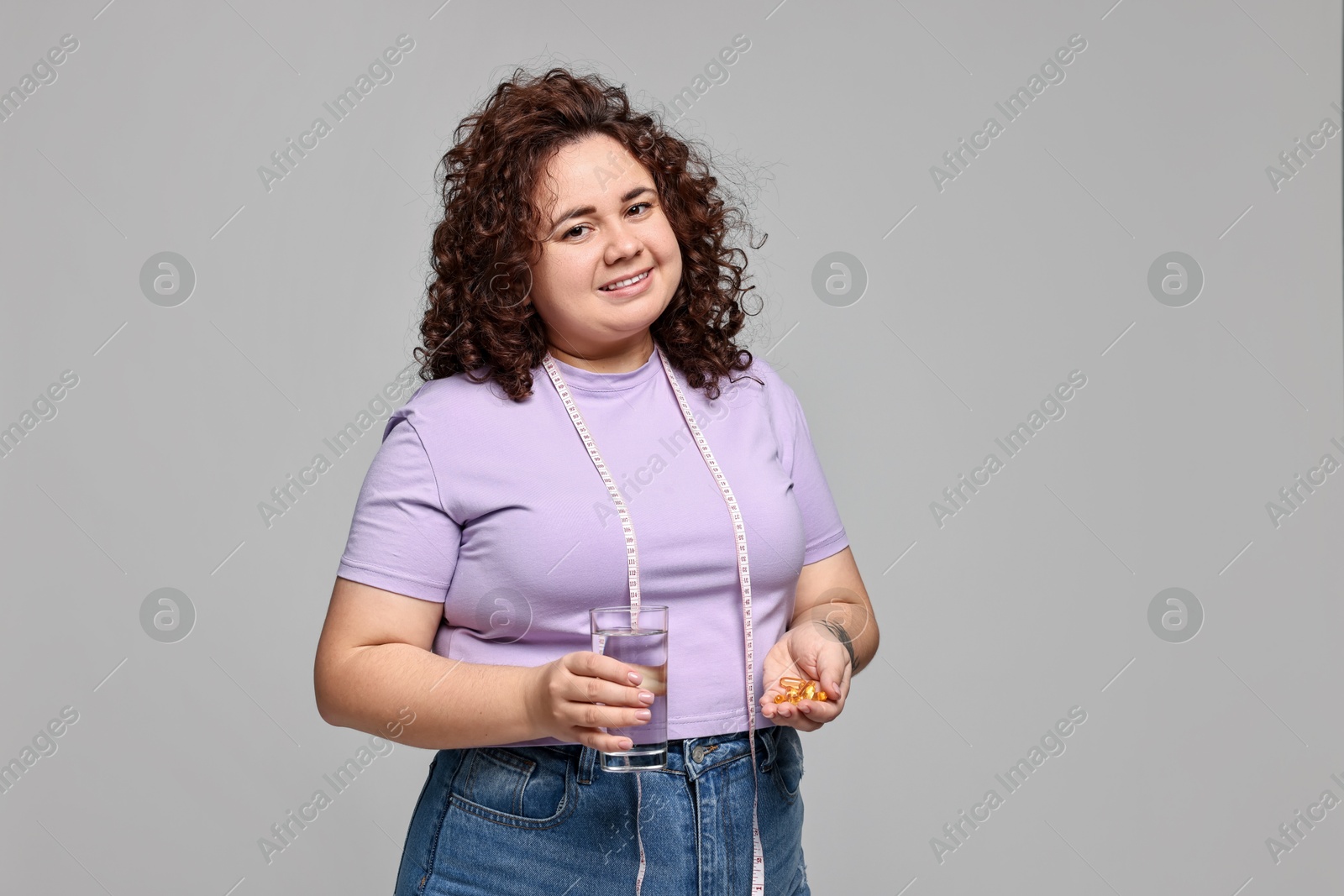 Photo of Happy plus size woman with pile of weight loss supplements and glass of water on grey background