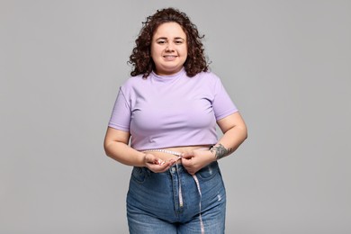 Happy plus size woman measuring waist with tape and holding pile of weight loss supplements on grey background