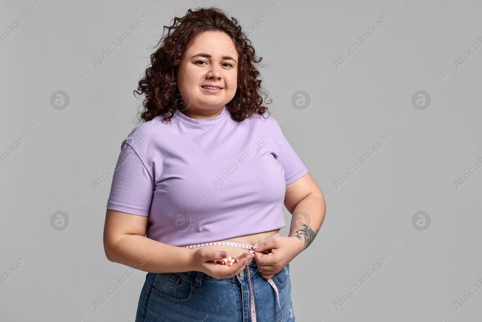 Photo of Happy plus size woman measuring waist with tape and holding pile of weight loss supplements on grey background