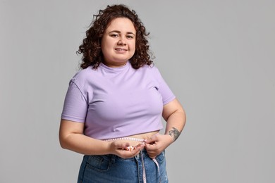 Photo of Happy plus size woman measuring waist with tape and holding pile of weight loss supplements on grey background