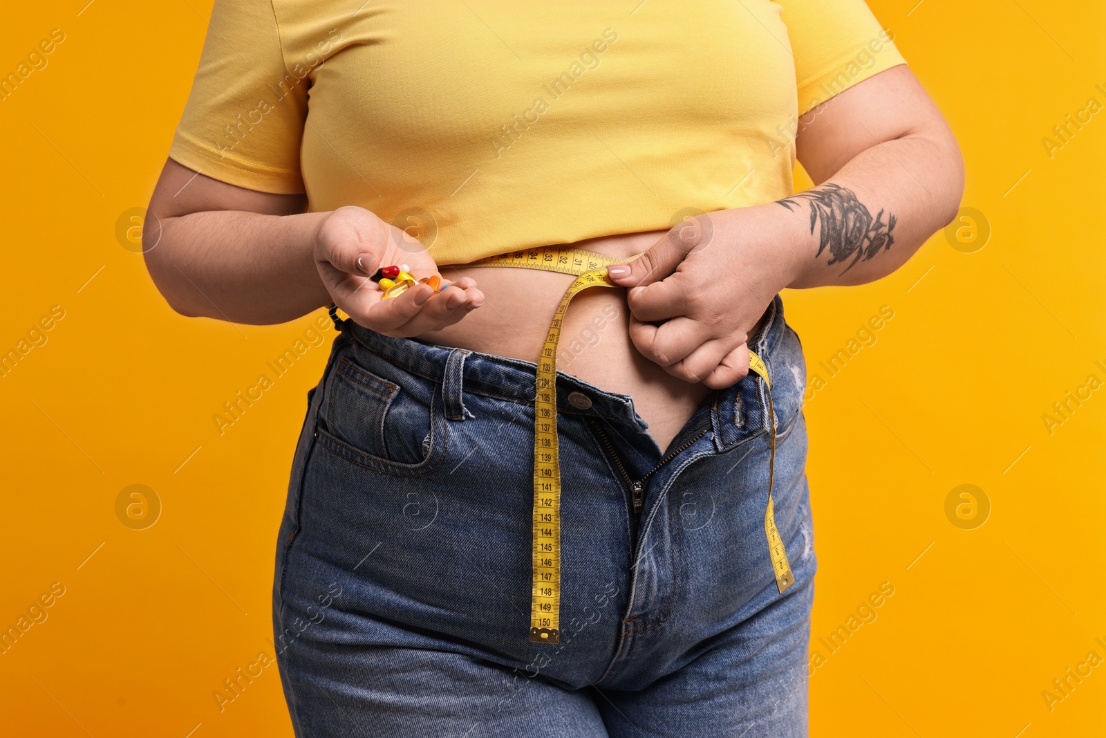 Photo of Plus size woman measuring waist with tape and holding pile of weight loss supplements on orange background, closeup