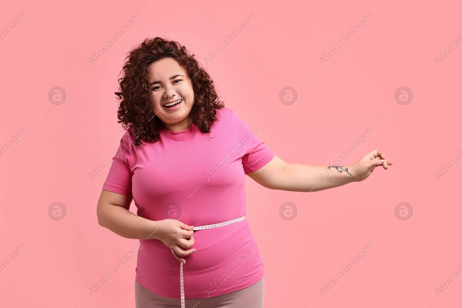 Photo of Weight loss. Happy plus size woman measuring waist with tape on pink background