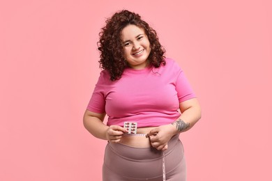 Photo of Happy plus size woman measuring waist with tape and holding blister of weight loss supplements on pink background