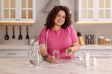 Photo of Happy plus size woman with weight loss supplements and glass of water at marble countertop in kitchen