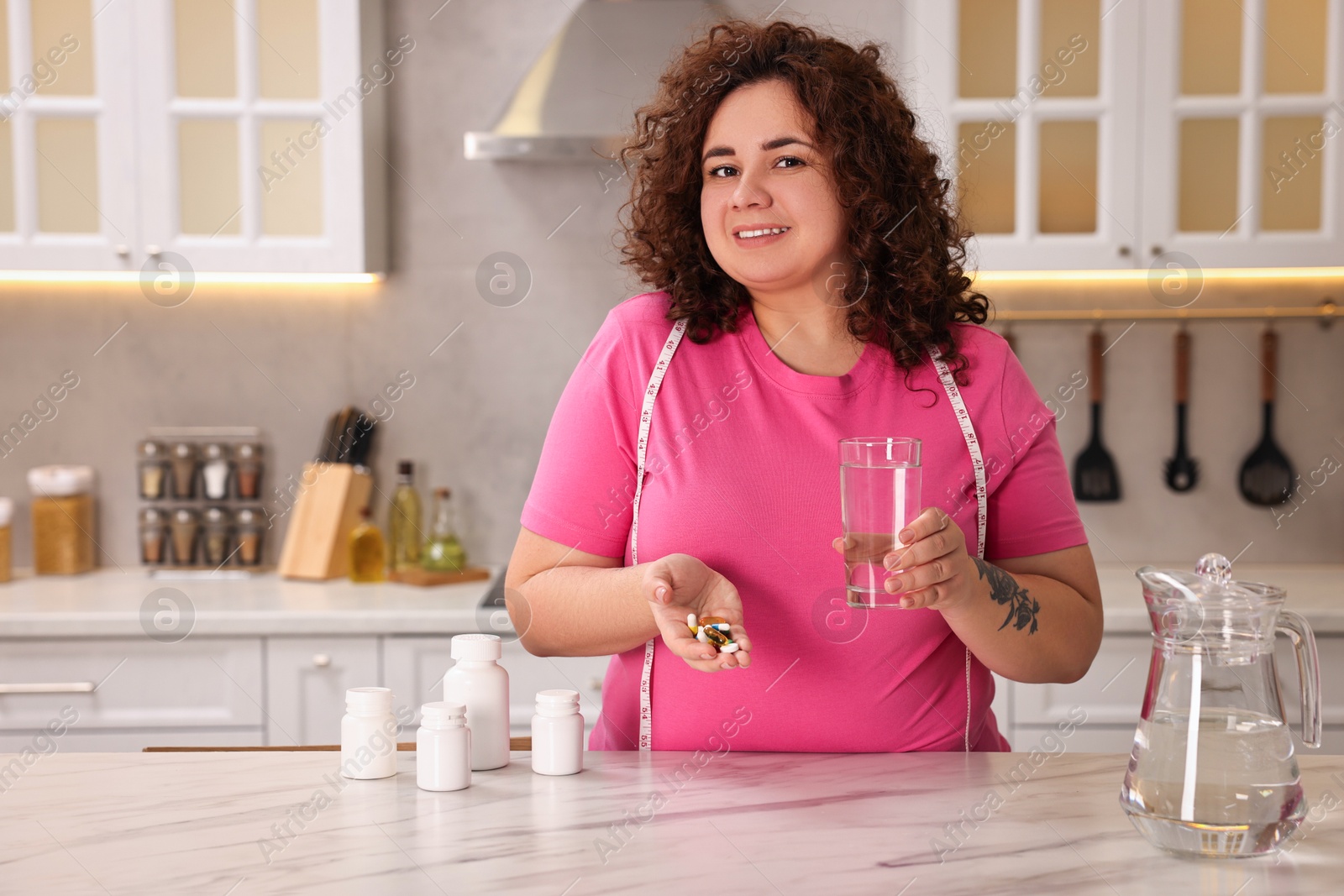 Photo of Happy plus size woman with weight loss supplements and glass of water at marble countertop in kitchen