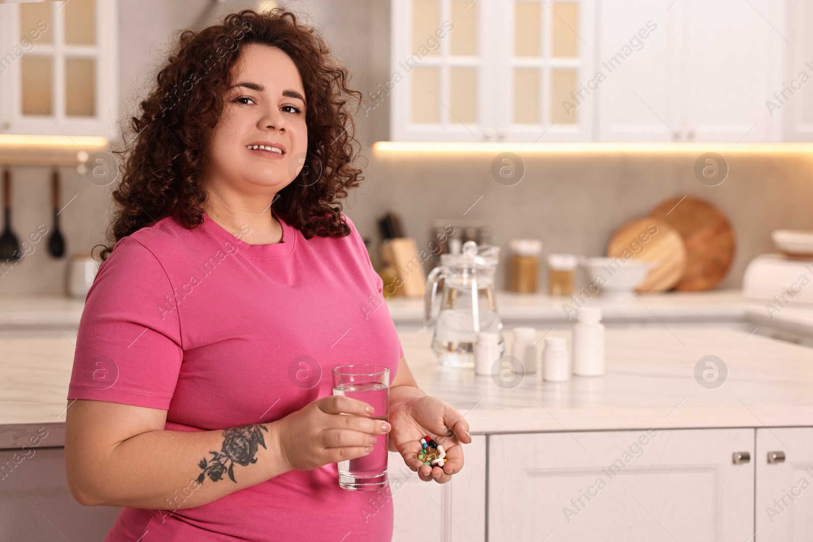 Photo of Happy plus size woman holding pile of weight loss supplements and glass with water in kitchen. Space for text