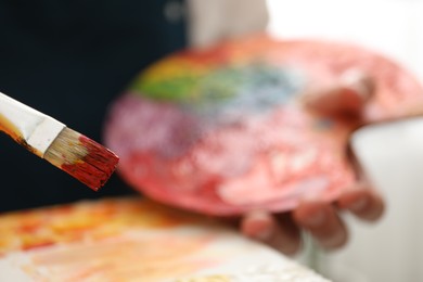 Man with palette and paintbrush drawing picture indoors, selective focus