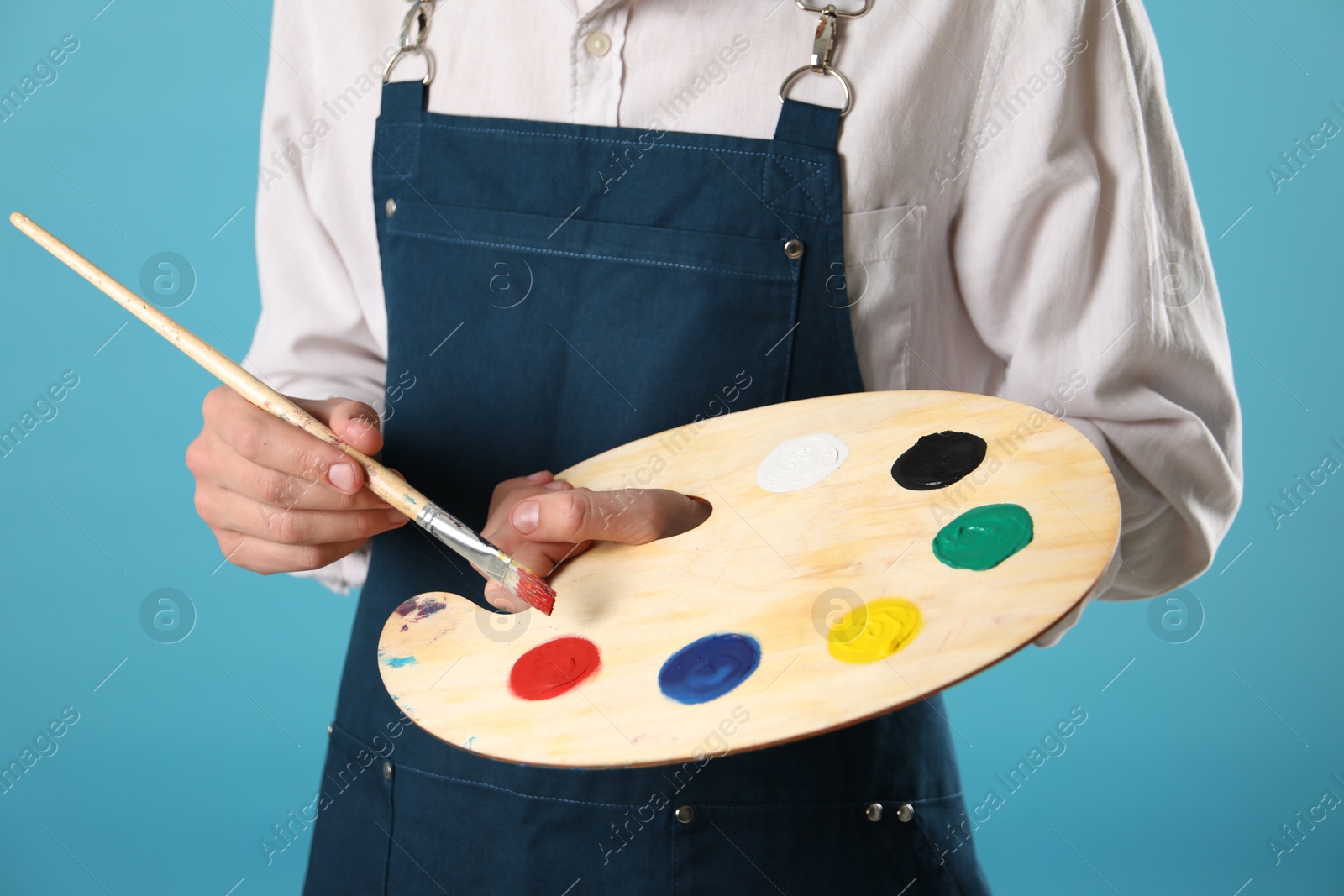 Photo of Man with wooden palette and paintbrush on light blue background, closeup