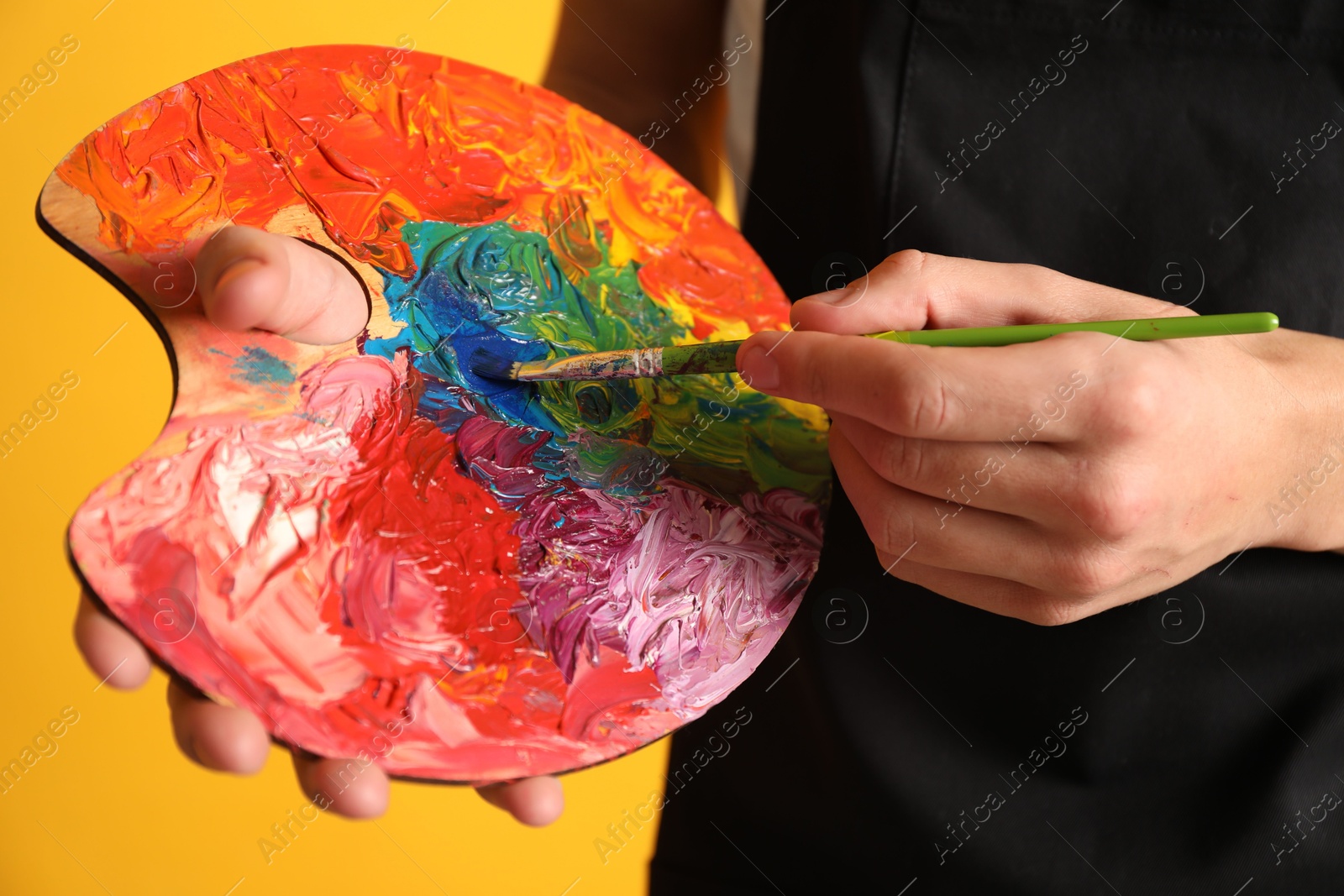 Photo of Man with paintbrush mixing paints on palette against orange background, closeup