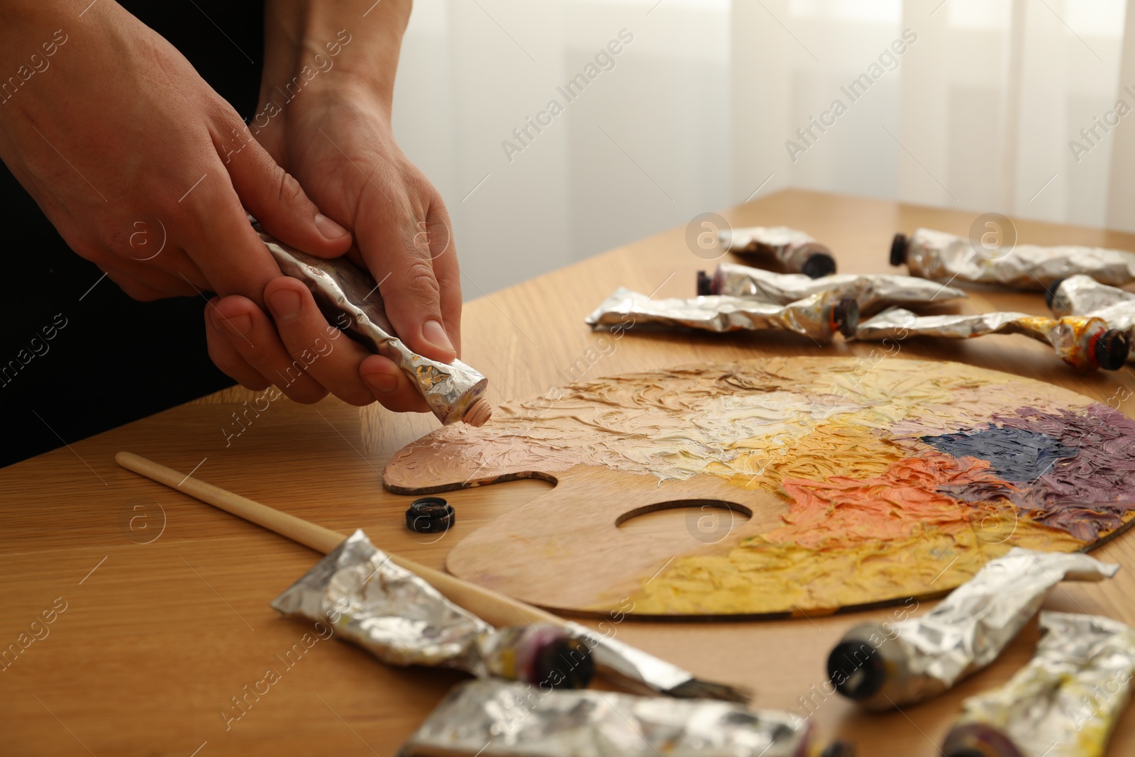 Photo of Man squeezing paint from tube onto palette at wooden table indoors, closeup