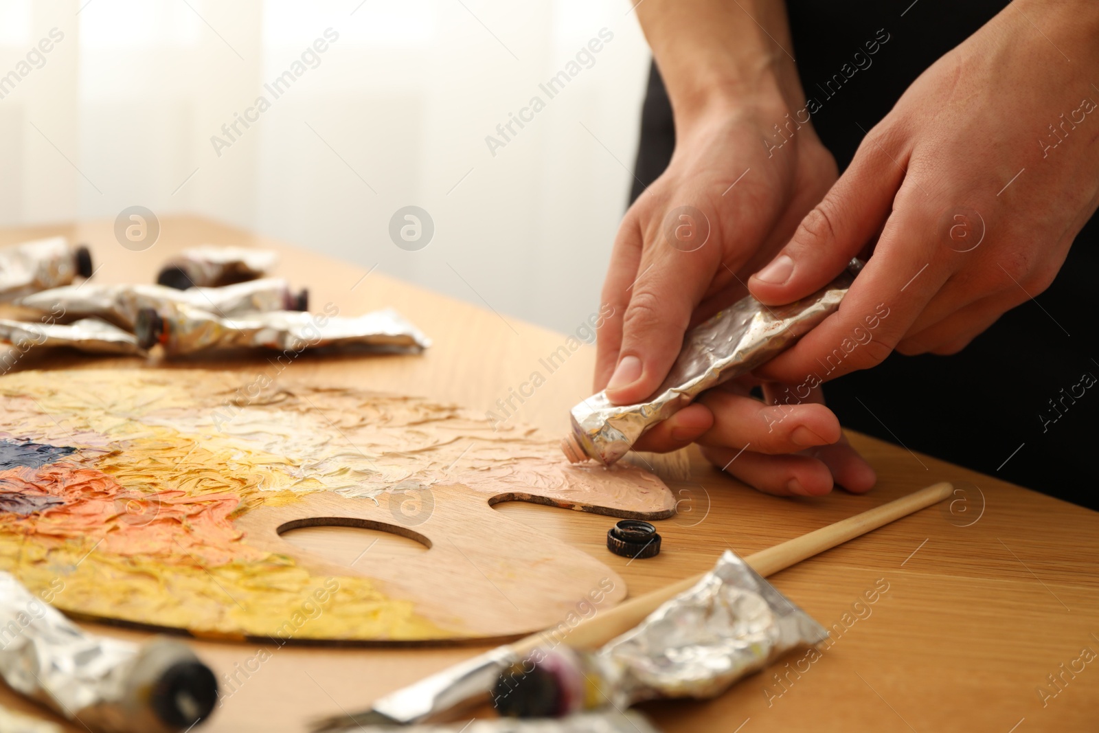 Photo of Man squeezing paint from tube onto palette at wooden table indoors, closeup