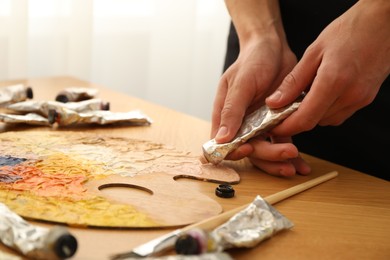 Man squeezing paint from tube onto palette at wooden table indoors, closeup