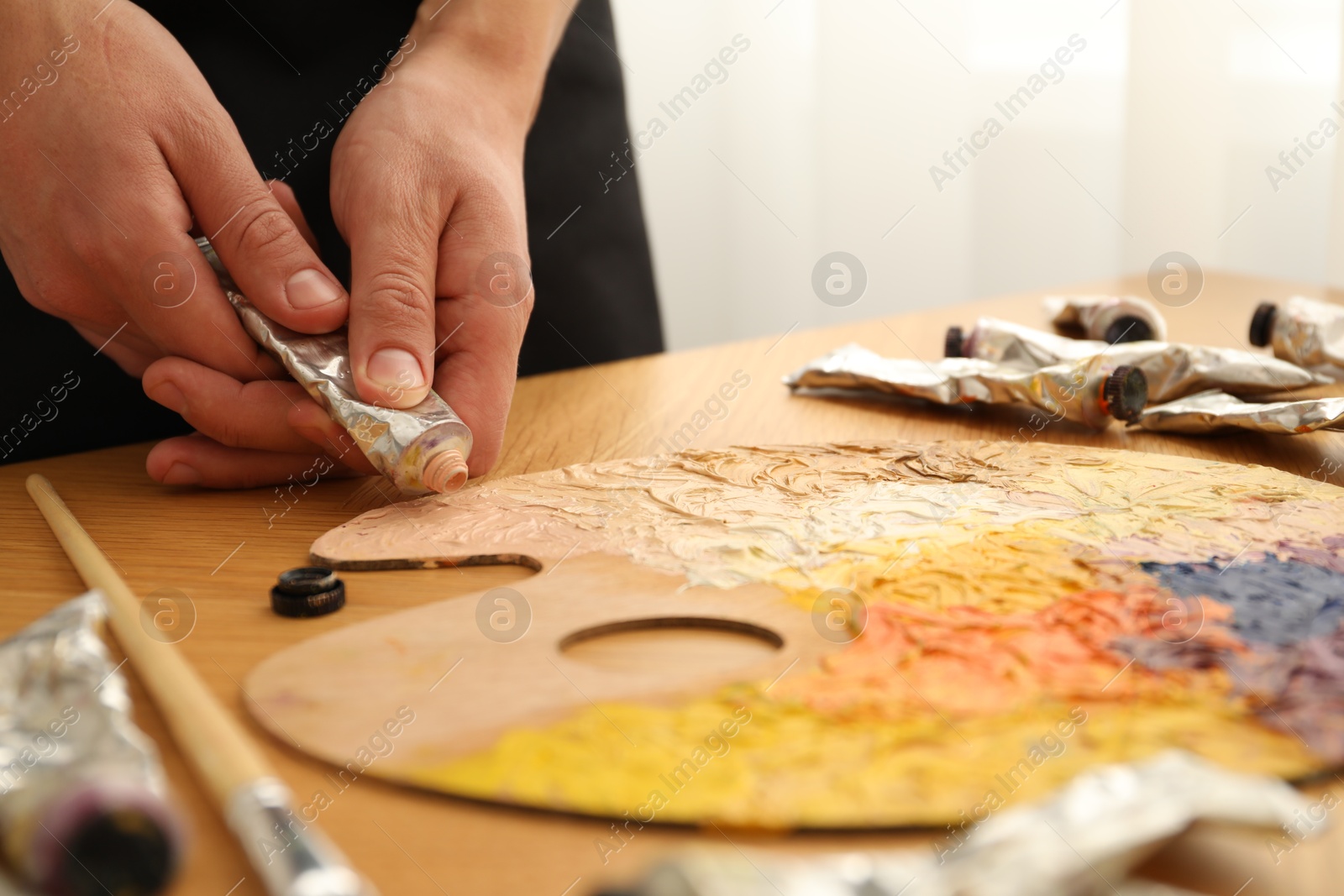 Photo of Man squeezing paint from tube onto palette at wooden table indoors, closeup