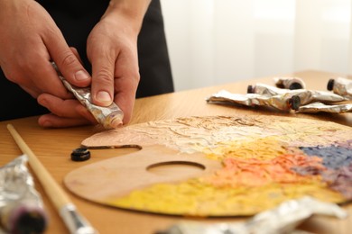 Man squeezing paint from tube onto palette at wooden table indoors, closeup