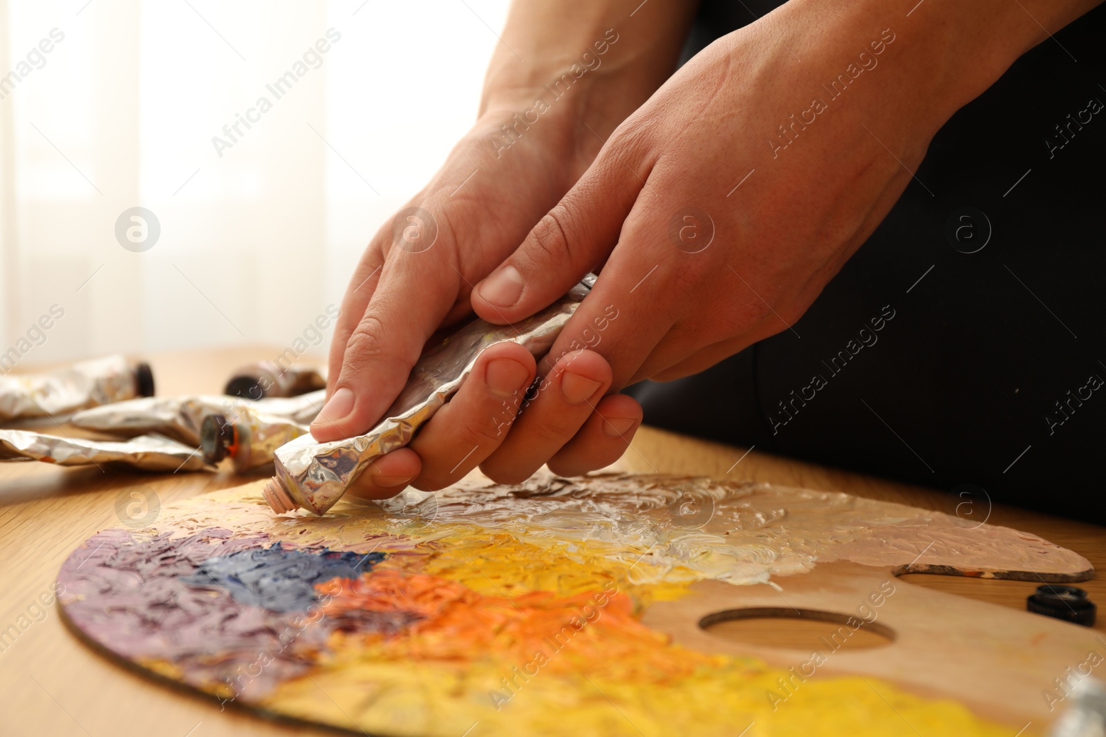 Photo of Man squeezing paint from tube onto palette at wooden table indoors, closeup