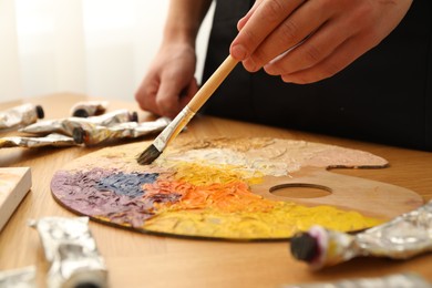 Photo of Man with paintbrush mixing paints on palette at wooden table indoors, closeup