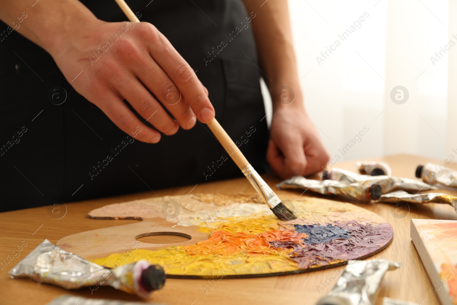 Photo of Man with paintbrush mixing paints on palette at wooden table indoors, closeup