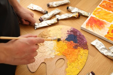 Photo of Man with paintbrush mixing paints on palette at wooden table, closeup