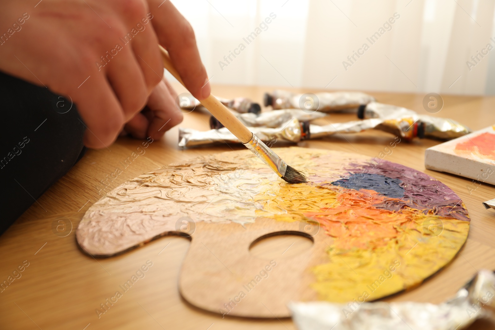 Photo of Man with paintbrush mixing paints on palette at wooden table indoors, closeup