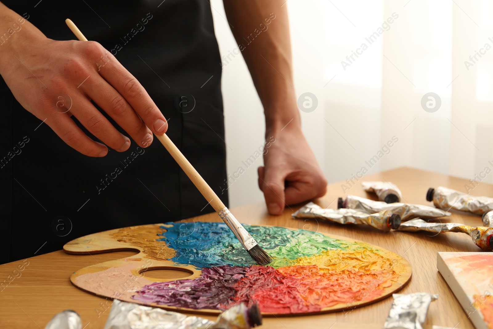 Photo of Man with paintbrush mixing paints on palette at wooden table indoors, closeup