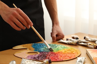 Photo of Man with paintbrush mixing paints on palette at wooden table indoors, closeup