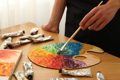 Photo of Man with paintbrush mixing paints on palette at wooden table indoors, closeup