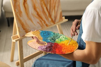 Man with palette and paintbrush drawing picture indoors, closeup